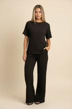 A woman takes a mirror selfie wearing a matching black textured short-sleeve top and wide-leg pants set against a plain white wall.