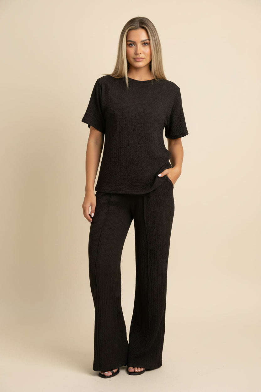 A woman takes a mirror selfie wearing a matching black textured short-sleeve top and wide-leg pants set against a plain white wall.